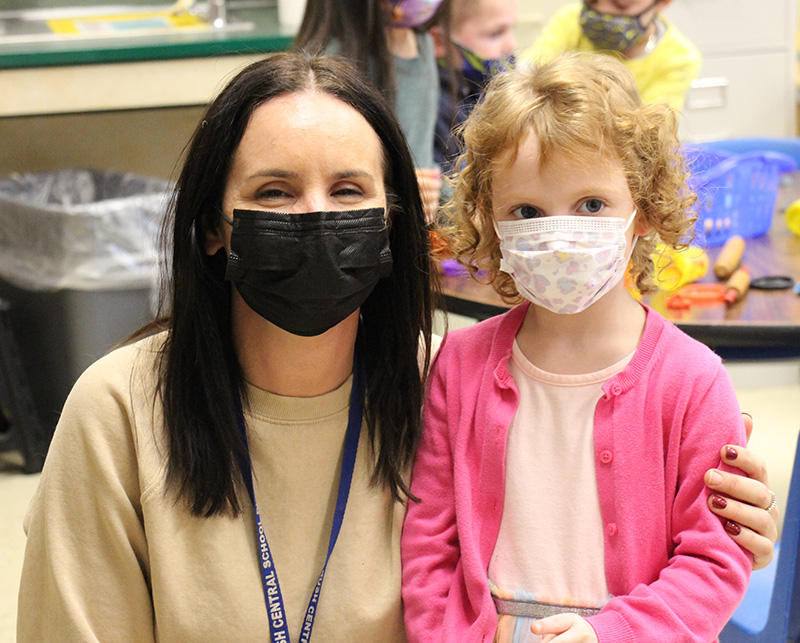 A woman with long dark hair, wearing a black mask and tan sweater, squats down next to a little girl with short blonde hair, wearing a pink sweater and light-colored mask.