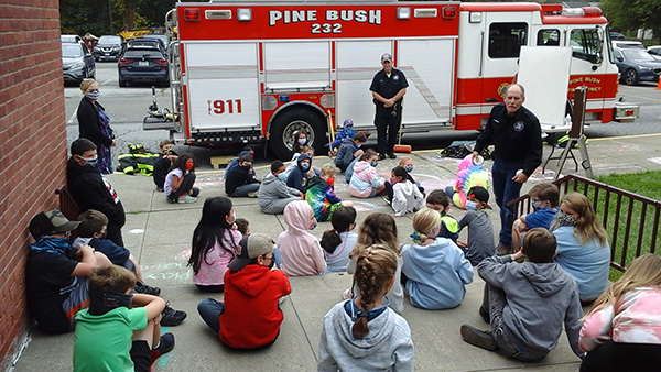 A firefighter dressed in navy blue stands in front of a red firetruck. There is an entire class of smaller elementary students sitting on the concrete, spaced apart from each other, facing him and listening.