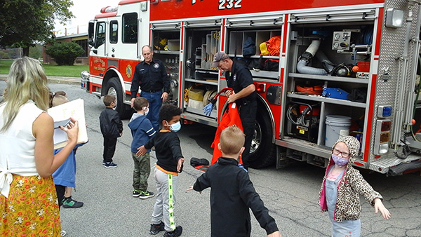 Two firefighters dressed in navy blue stand in front of a fire truck, which has its sides up and equipment showing. There are five elementary students standing at a distance from each other watching the men.