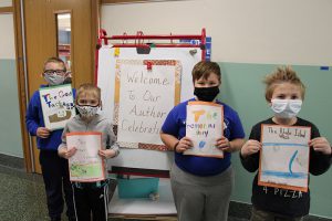 A large signs says Welcome to our Author Celebration. In front of it are four students , all masked, holding their handwritten books in front of them.