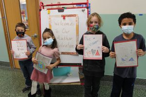 A large signs says Welcome to our Author Celebration. In front of it are four students , all masked, holding their handwritten books in front of them.