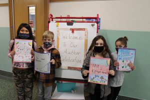 A large signs says Welcome to our Author Celebration. In front of it are four students , all masked, holding their handwritten books in front of them.