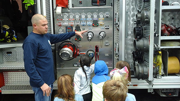 A man in a blue shirt points to gadgets on a fire truck as five elementary students watch. One girl with long dark hair points to something on the truck.