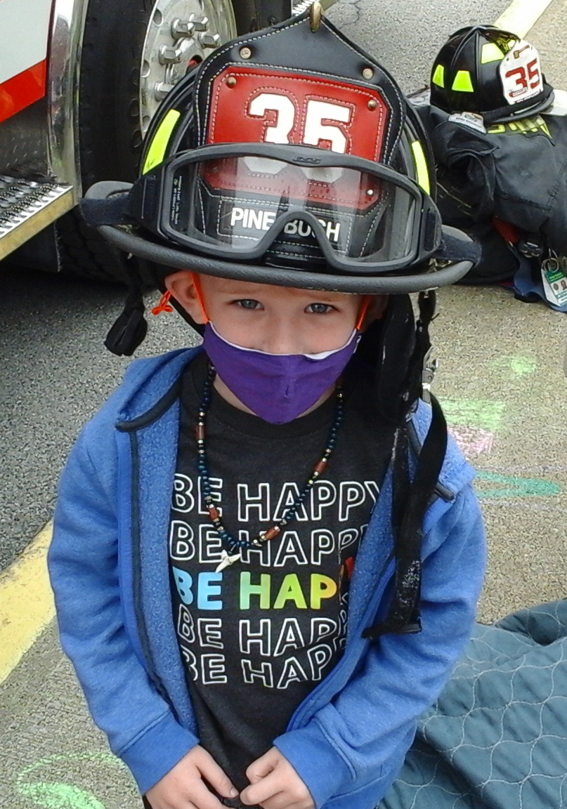 A small elementary student, wearing a blue mask and a shirt that says Be Happy and a blue jacket, wears a firefighter helmet with the number 35 on it