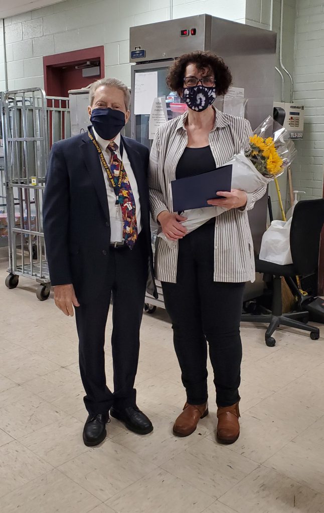 A man stands on the left wearing a suit and tie and a black mask. A woman on the right is holding yellow flowers. She is wearing black pants and shirt with a light printed shirt over it.