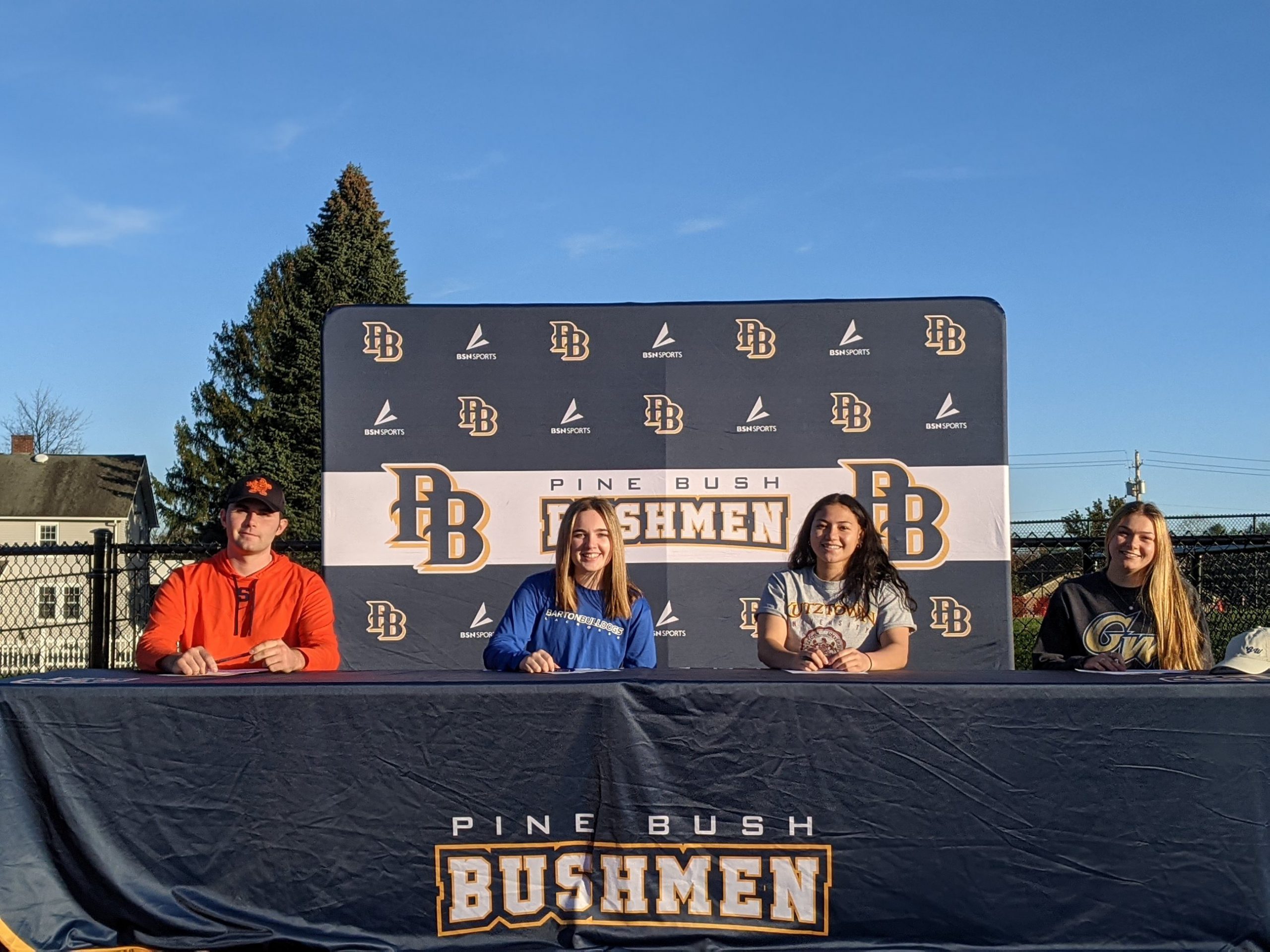 A bright blue sky in the background. Four high school seniors sitting at a table covered in a blue tablecloth that says Pine Bush Bushmen. Behind them is a background the PB on it and Pine Bush Bushmen in blue and gold. From left, a young man wearing an orange shirt and a Syracuse hat; a young woman with long blonde hair wearing a blue shirt that says Barton College, A girl with long dark hair wearing agray shirt that says Kutztown University and a girl on the end with long blonde hair wearing a blue jersey that says George Washington University. They are all smiling.