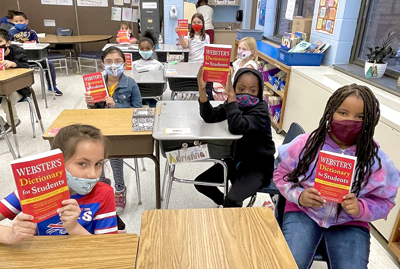 Seven elementary students sitting at desks holding up red dictionaries. All are wearing masks.