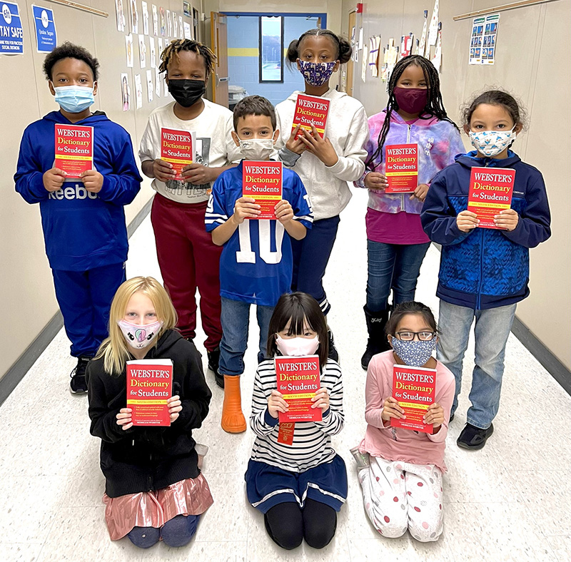 Three third grade students kneel in front and six stand in back. All are wearing masks and holding up red dictionaries.