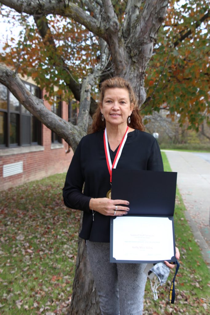 A woman stands outside by a tree. She is holding a certificate and wearing a medal around her neck. She is smiling.