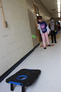 A girl wearing pink pants and a blue and pink shirt tosses a beanbag at a cornhole table.