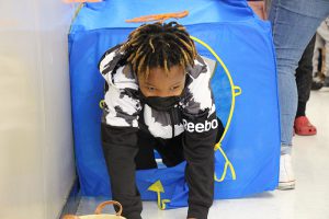 An elementary students wearing a black and white shirt and black mask comes through a hole in a blue box, part of an obstacle course.