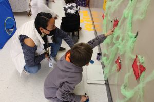 A young boy is sitting on the floor and pointing to paper on the wall, which also has green material that looks like spider webs. A woman is behind him squating and pointing too.