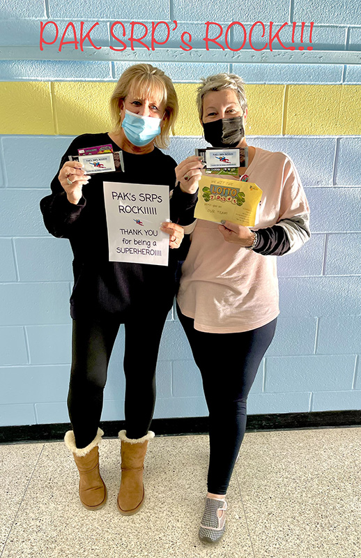 Two women stand in front of a blue and yellow wall. They are both wearing masks and holding certificates.