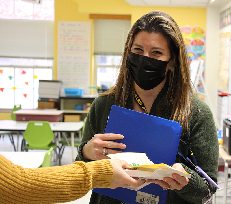 A woman with long blonde hair, wearing a sweater and a black mask, accepts a gift from a woman not on camera. She smiles behind the mask.