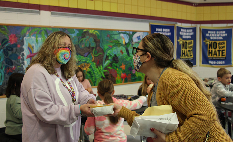 A woman with blonde hair pulled back in a ponytail, wearing a mask and a gold sweater, hands a gift to a woman with shoulder-length blonde hair, wearing a tie-dye mask and glasses. There are kids in the background.