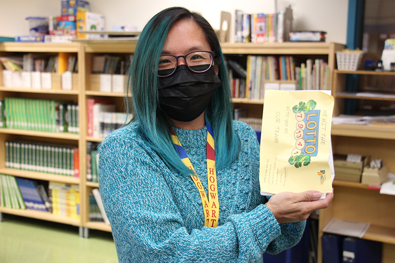 A woman wearing a blue sweater and black mask, with longish hair tinged in blue and glasses, holds up a gift she received.