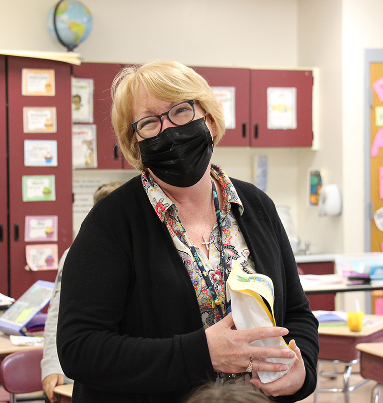 A woman with short blonde hair, wearing glasses, black mask and black sweater holds a gift. She has her head tilted to the left and is smiling.