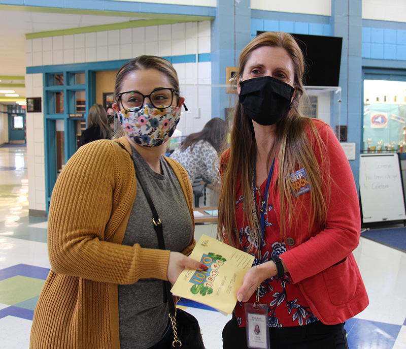 A woman with blonde hair pulled back, wearing glasses, a printed mask and a gold sweater, hands a gift to a woman with long blonde hair, wearing a black mask and rust colored shirt.