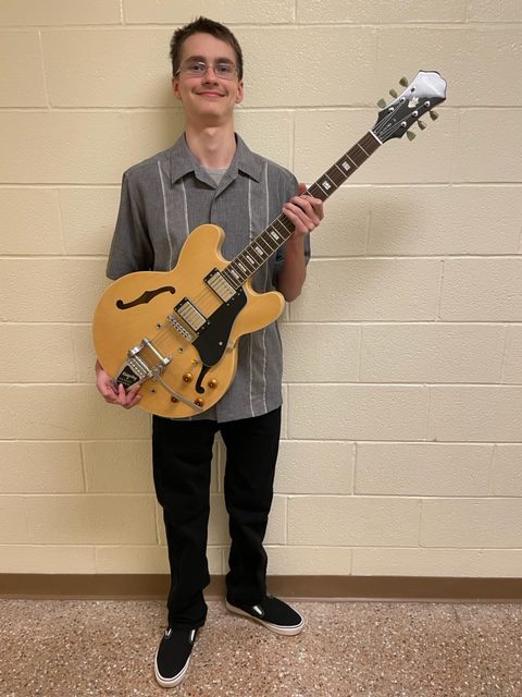 A high school boy dressed in a short-sleeve gray shirt and dark pants. He is smiling and holding a guitar.