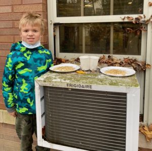 A boy in a gren and blue jacket stands in front of a building next to a window. There is an air conditioner in the window and on top of it are plates and cups with bird seed.