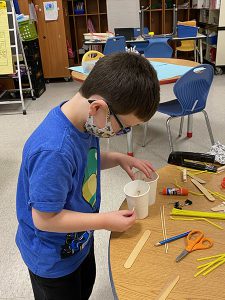 A boy wearing a blue shirt and glasses and a mask. He has dark hair and he isat a table making his bird feeder with cups. There are pencils, scissors and rulers on the table.