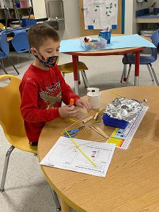 A boy in a red shirt wearing a mask sits at a table with supplies laid out. He has glue sticks, cups, paper.