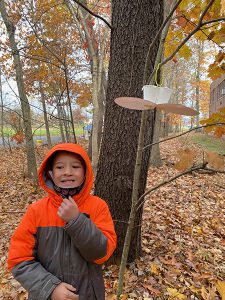 An elementary age boy in an orange and gray jacket stands smiling, in front of a tree where a birdfeeder is hanging.