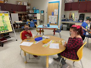 Two elementary students, a boy wearing red shirt and black mask, and a girl with a red and black checked shirt and dark mask, sit at a table making bird feeders. There are supplies on the table.