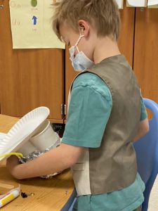 A boy wearing a green shirt and brown vest holds his birdfeeder, made of cups and paper plates. He is wearing a blue mask.