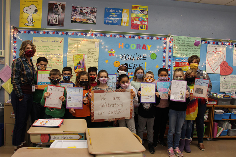 A class of third-grade students about 16 of them stand in front of the classroom holding their handmade books. On either side of the group is a teacher. All have masks on.