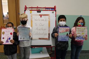 A large signs says Welcome to our Author Celebration. In front of it are four students , all masked, holding their handwritten books in front of them.