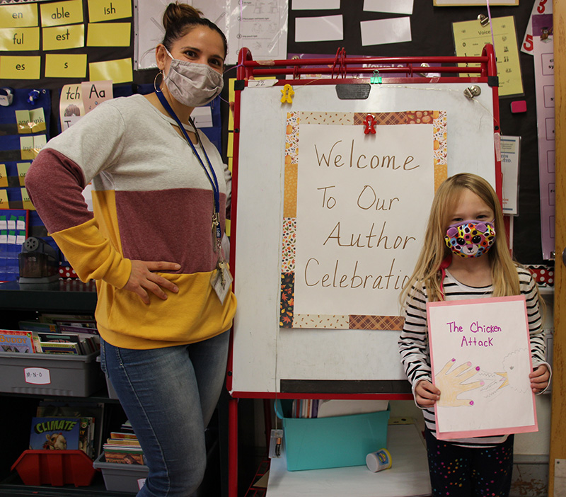 A large signs says Welcome to Our Author Celebration. on the left is a woman wearing a mask and a shirt that's white, brown and gold. On the right side is a girl with long blonde hair, wearing a mask holding her handwritten book.