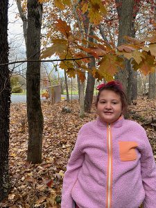 An elementary girl wearing a purple fleece jacket and a bow in her hair stands by a tree with red and brown leaves all around. There is a bird feeder hanging from the limb of the tree.