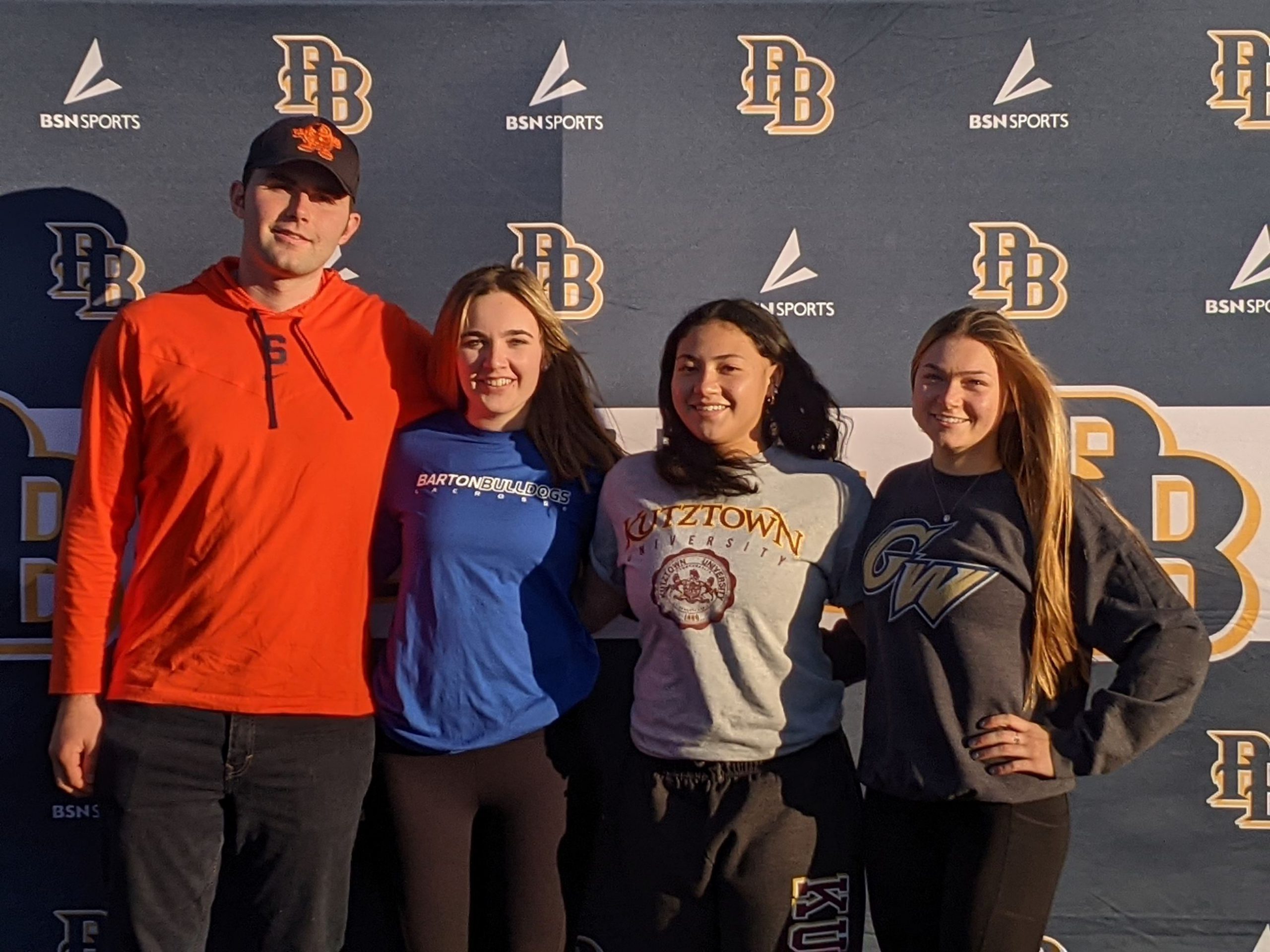 Four high school seniors standing arm in arm in front of a blue background with PB in From left, a young man wearing an orange shirt and a Syracuse hat; a young woman with long blonde hair wearing a blue shirt that says Barton College, A girl with long dark hair wearing agray shirt that says Kutztown University and a girl on the end with long blonde hair wearing a blue jersey that says George Washington University. They are all smiling.blue and gold on it. 