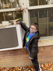 An elementary age boy, wearing a blue shirt and black jacket, glasses and a mask, reaches up to the cup and plate bird feeder he placed on top of an air conditioner that is in a window.