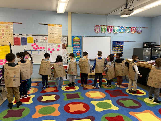 Twelve kindergarten students stand around a classroom, with their backs to the camera, all wearing brown paper vests with handdrawn pictures on it. There is one adult on the right . All are wearing face masks. They are standing on a multicolored rub with different shapes on it.