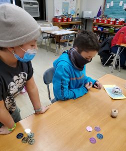 Two students are at a wooden table with a large wooden dreidel spinning. The boys are wearing masks and they are watching the dreidel. There are coins on the table.