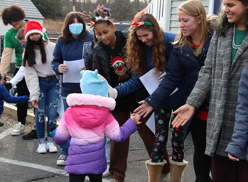 A line of middle school students stand with their hands out and down as little preschool kids walk by and give them high five.