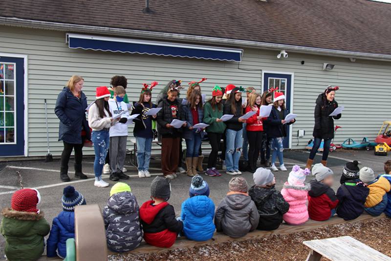 A group of about 15 middle school students with two adults stand in front of a building and sing christmas songs. Many are wearing festive hats and headbands. In front of them is a group of preschool kids sitting on a railroad tie. All are dressed for winter.