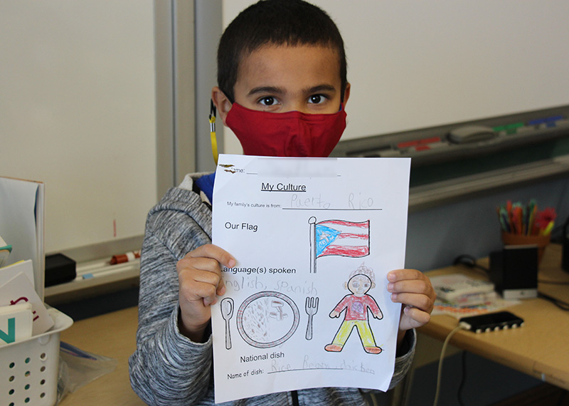 An elementary-age boy with short dark hair wearing a red mask and gray shirt holds up a piece of paper with a flag of Puerto Rico on it.