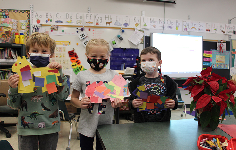 Three kindergarten kids - a boy on the left, a girl in center, and a boy on right - all hold cut outs of a horse pinata. They are decorated with multiple pieces of colorful pieces of paper glued on them. The children are all wearing masks.