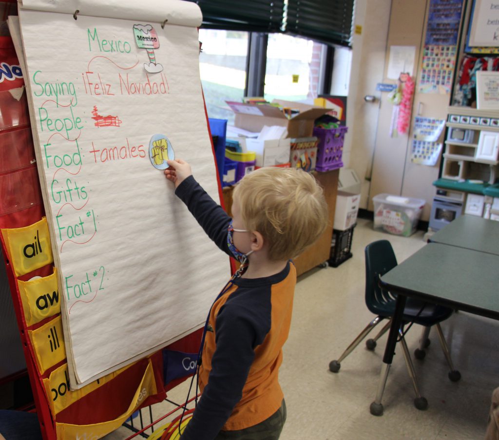 A boy with blond hair, wearing a black and orange shirt puts a sticker on a large poster that says Mexico and lists holiday traditions.