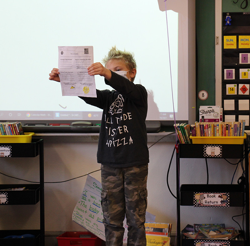 An elementary-age boy with blonde hair holds up a piece of paper with information about his heritage. He is wearing a dark shirt and a mask.