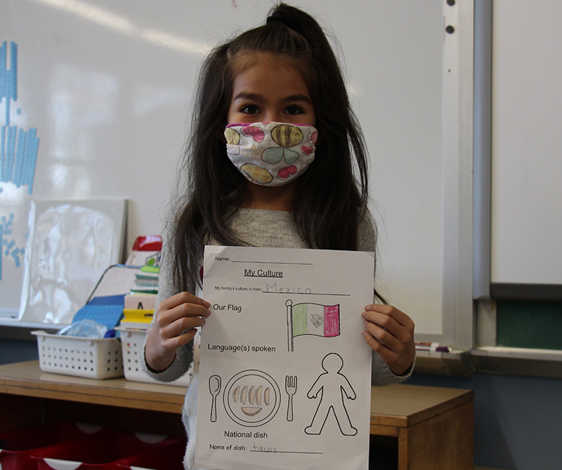 A small elementary school girl with long dark hair, wearing a light-colored print mask. She is holding a piece of paper showing she is from Mexico.