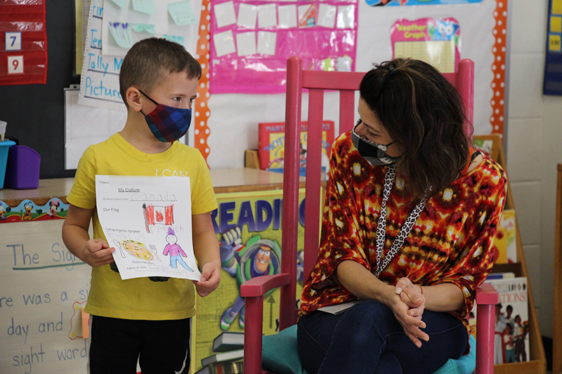 A woman with dark hair, a print shirt  sits on a chair looking at a little boy who is explaining to her where his family is from. He is wearing a yellow shirt and dark mask, holding a piece of paper with the flag of Canada.