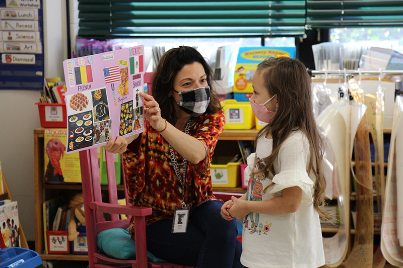 A woman with dark hair and mask, and a print shirt holds up a poster that has several flats on it as well as pictures of food. A little girl is looking at the poster and telling her about what's on it. The girl has long brown hair and is wearing a mask and white shirt.