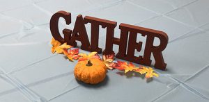 A wooden cutout that says Gather. In front of it are paper leaves in fall colors and a small pumpkin.