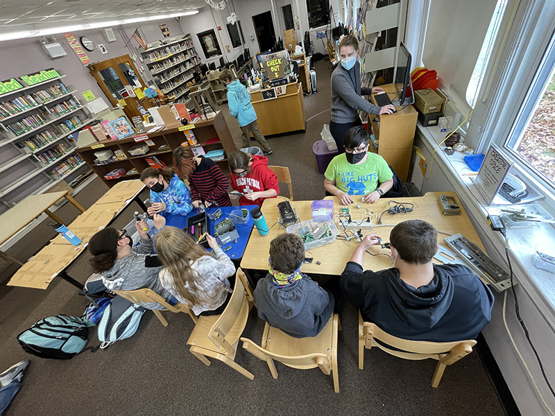 Photo is taken from above. Middle schools students -  about 8 of them, are sitting at tables working on their  sculptures using parts of old electronics.