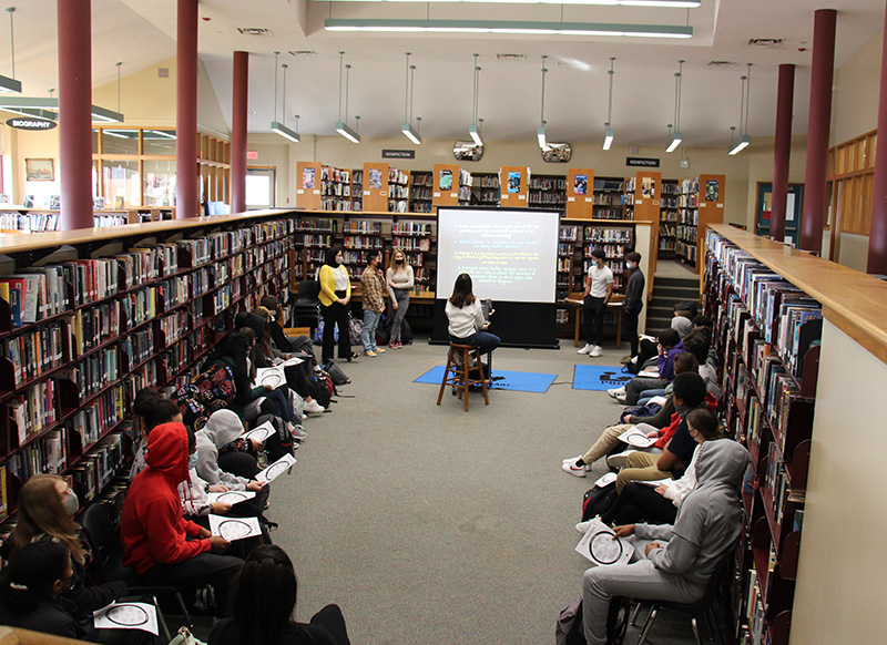 A smaller part of a library with students around the perimeter. In the front is a large screen showing information from a projector. There are high school students at the front running the projector and talking. All  are wearing masks.