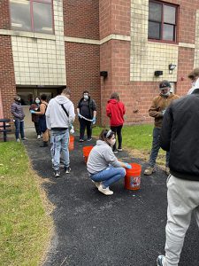 high school students are outside on a paved walkway. There are orange buckets on the walkway where the students are crouching to put their shirts in for tie dye.